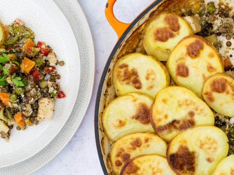 A lentil hotpot with Quorn Meat Free Pieces and mixed vegetables topped with sliced potatoes in a shallow orange Dutch oven with one portion removed and plated on the side.
