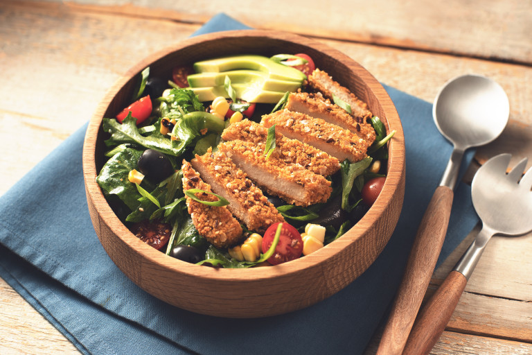 A mixed green salad with corn, cherry tomatoes, and olives topped with sliced avocado and a sliced Quorn Vegan Meatless Chipotle Cutlet in a wooden bowl with wood-handled utensils to the right.