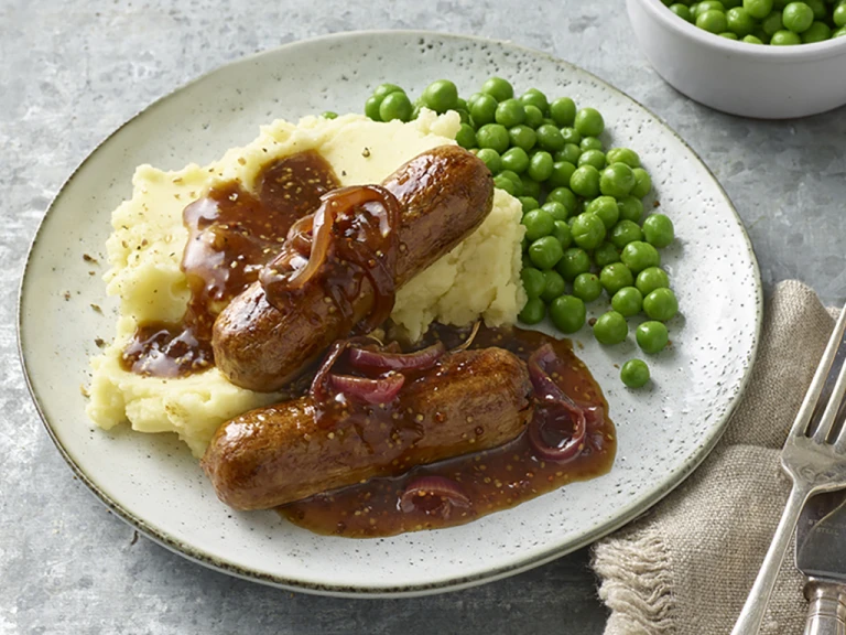 Plate containing sausages with red onion gravy upon garlic mash and peas alongside a cutlery and an extra side of peas