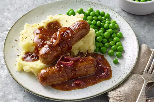 Plate containing sausages with red onion gravy upon garlic mash and peas alongside a cutlery and an extra side of peas
