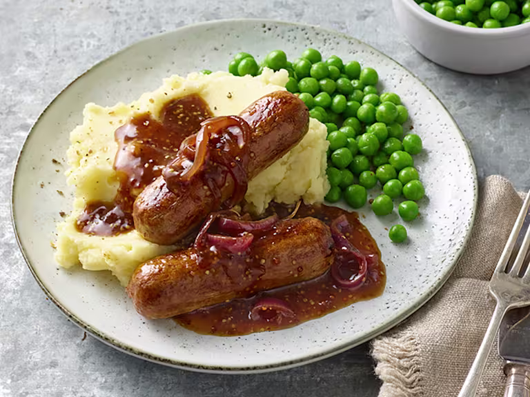 Plate containing sausages with red onion gravy upon garlic mash and peas alongside a cutlery and an extra side of peas