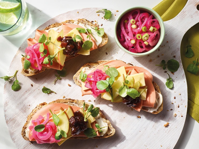 Three open-faced vegan ploughman's sandwiches topped with Quorn Vegan Smoky Ham Free Slices, Violife Epic Mature Cheddar Block slices, apple slices, ploughman's relish, pea shoots, and pickled onions arranged on a board.