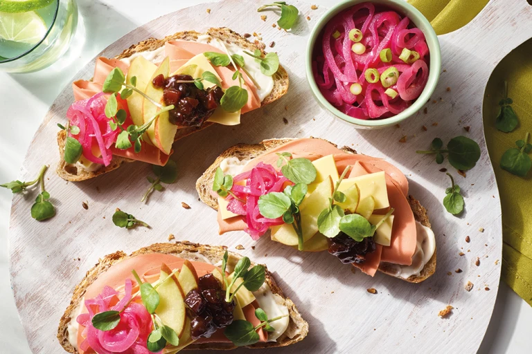 Three open-faced vegan ploughman's sandwiches topped with Quorn Vegan Smoky Ham Free Slices, Violife Epic Mature Cheddar Block slices, apple slices, ploughman's relish, pea shoots, and pickled onions arranged on a board.