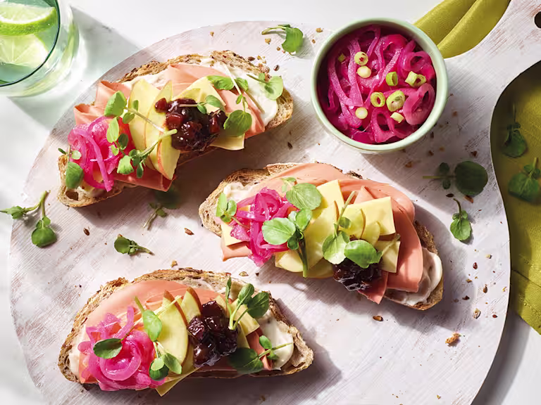 Three open-faced vegan ploughman's sandwiches topped with Quorn Vegan Smoky Ham Free Slices, Violife Epic Mature Cheddar Block slices, apple slices, ploughman's relish, pea shoots, and pickled onions arranged on a board.