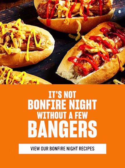 Hot dogs with onions, mustard, and ketchup on a wooden board, served with sweet potato fries and corn salsa. Text reads: “It’s not Bonfire Night without a few bangers” and “View our Bonfire Night recipes.”