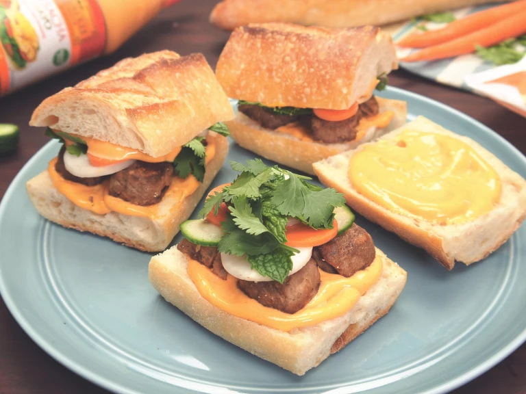 Two small banh mi sandwiches with pickled carrots and daikon, cucumber, Quorn Meatballs, cilantro, mint, and sriracha mayo on a baguette atop a blue plate.