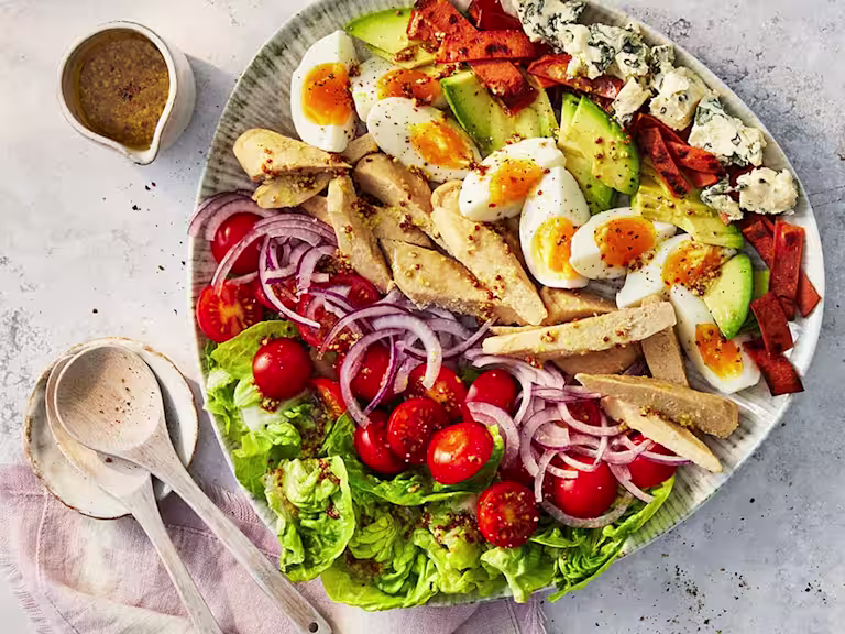 Layered salad on a ceramic plate with lettuce, chopped cherry tomatoes, sliced red onion, Quorn Roast Mini Fillets, sliced boiled egg, sliced avocado, grilled red pepper and cubes of blue cheese.