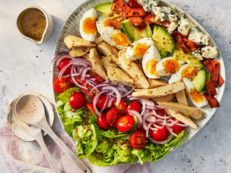 Layered salad on a ceramic plate with lettuce, chopped cherry tomatoes, sliced red onion, Quorn Roast Mini Fillets, sliced boiled egg, sliced avocado, grilled red pepper and cubes of blue cheese.