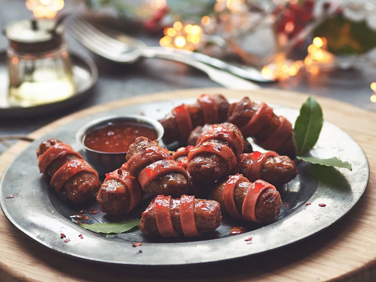 Vegetarian Pigs in Blankets made with Quorn Cocktail Sausage wrapped in Quorn Vegan Ham Slices on a plate with bay leaves and tomato sauce.