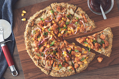 Quorn Strips, cheese, corn, red onion, and green onions atop a cauliflower crust on a wooden board with barbecue sauce and a pizza slicer on either side.