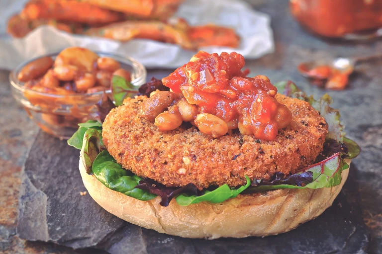 A Quorn Vegan Meatless Spicy Patty on the bottom of a burger bun topped with mixed greens, spicy beans, and a tomato relish on a slate tray with a small glass bowl of beans in the background.