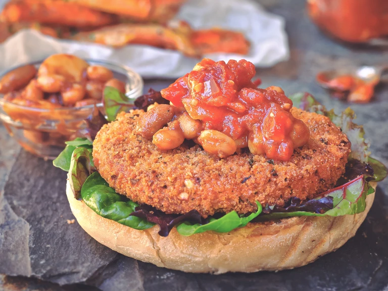 A Quorn Vegan Meatless Spicy Patty on the bottom of a burger bun topped with mixed greens, spicy beans, and a tomato relish on a slate tray with a small glass bowl of beans in the background.