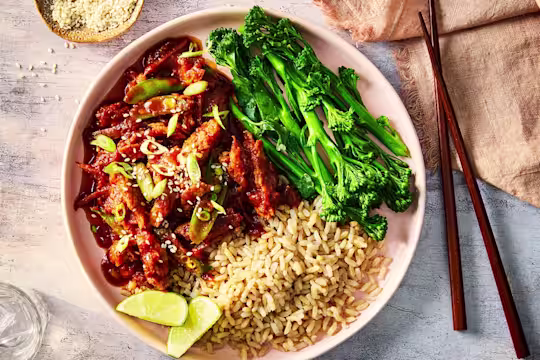 Honey and garlic chicken served with rice, tenderstem broccoli and lime wedges in a white dish with wood chopsticks.
