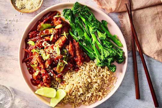 Honey and garlic chicken served with rice, tenderstem broccoli and lime wedges in a white dish with wood chopsticks.