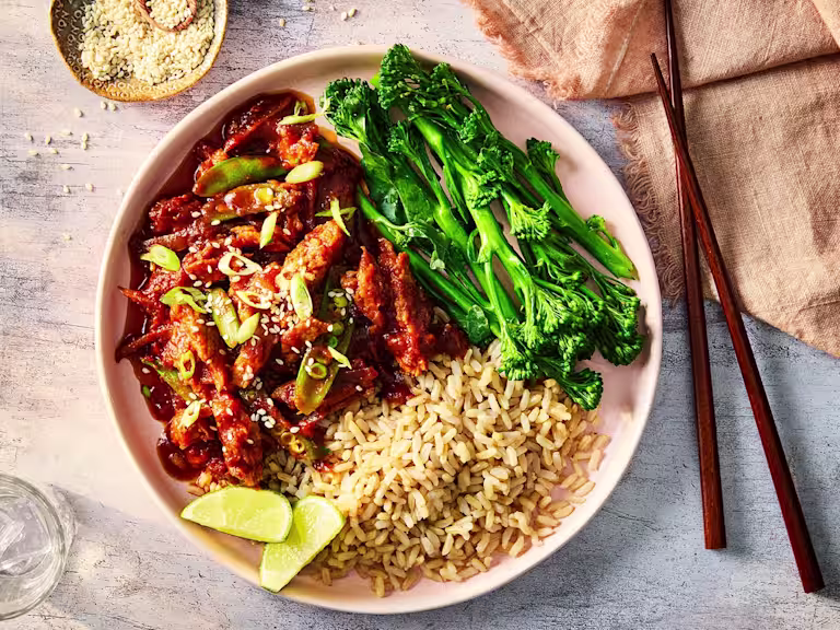Honey and garlic chicken served with rice, tenderstem broccoli and lime wedges in a white dish with wood chopsticks.