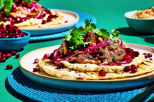 Flatbread topped with Quorn Vegan Pieces in Turkish spices, hummus, pickled red onion, pomegranate, coriander, and honey & lemon dressing on a blue placemat on a jade green backdrop.