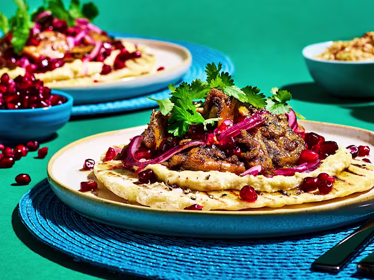 Flatbread topped with Quorn Vegan Pieces in Turkish spices, hummus, pickled red onion, pomegranate, coriander, and honey & lemon dressing on a blue placemat on a jade green backdrop.