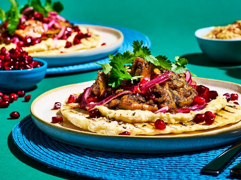 Flatbread topped with Quorn Vegan Pieces in Turkish spices, hummus, pickled red onion, pomegranate, coriander, and honey & lemon dressing on a blue placemat on a jade green backdrop.