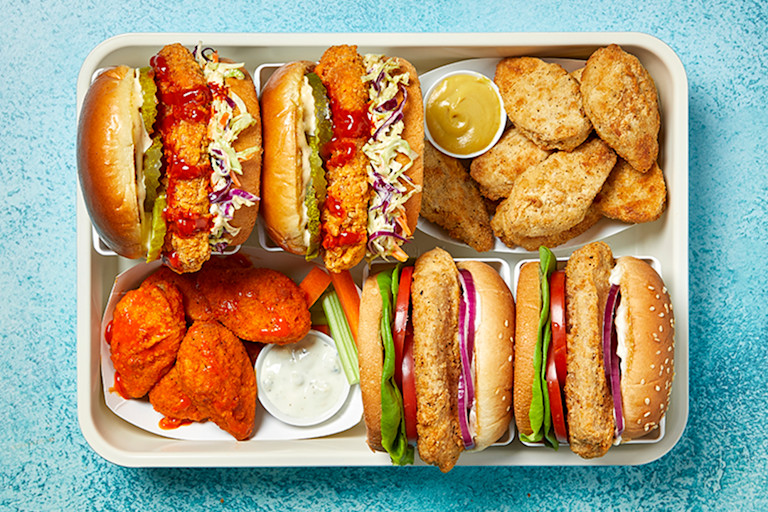 A burger, a chili cheese dog, nuggets in a sriracha glaze with blue cheese dressing on the side, a fried snack, carrot and red cabbage slaw, and sweet potato fries are arranged on a table with ramekins of sauces in between.