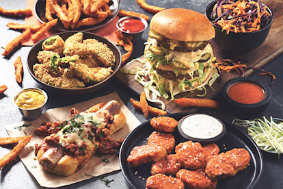 A burger, a chili cheese dog, nuggets in a sriracha glaze with blue cheese dressing on the side, a fried snack, carrot and red cabbage slaw, and sweet potato fries are arranged on a table with ramekins of sauces in between.