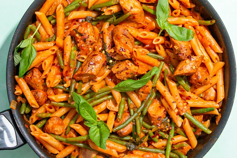: Quorn Sausage Arrabiata pasta in a pan with basil leaves and green beans