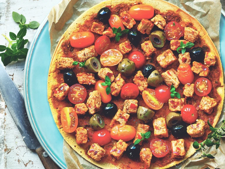 A socca pancake topped with black and green olives, grape tomatoes, microbasil, and Quorn Pieces on a piece of parchment atop a Tiffany blue plate with a knife to the left.