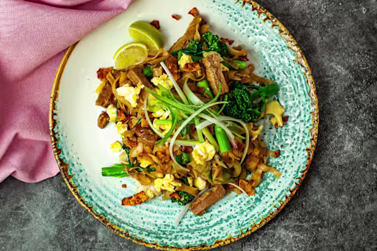 Pad See Ew served on a glazed plate with Quorn Steak Strips, tenderstem broccoli and slices of lemon with a pink napkin top left.
