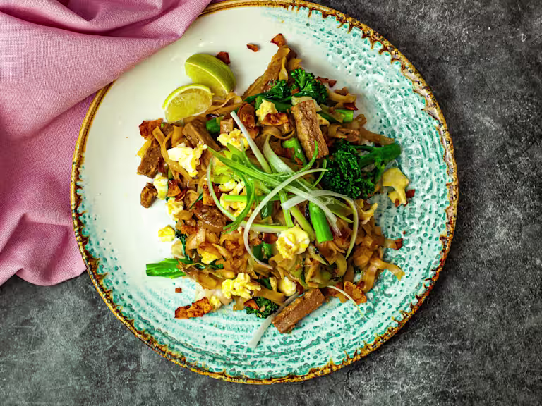 Pad See Ew served on a glazed plate with Quorn Steak Strips, tenderstem broccoli and slices of lemon with a pink napkin top left.