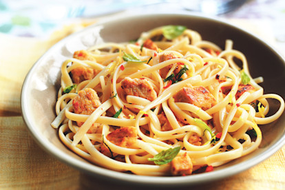 A taupe bowl of linguine topped with Quorn Pieces, chili flakes, and basil on an ochre tablecloth.