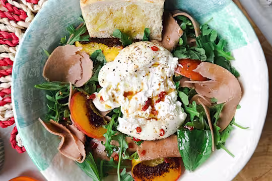 Ceramic bowl on a woven placemat with salad with rocket and basil, burrata cheese, grilled peaches, Quorn Vegetarian Ham Slices and toasted bread.