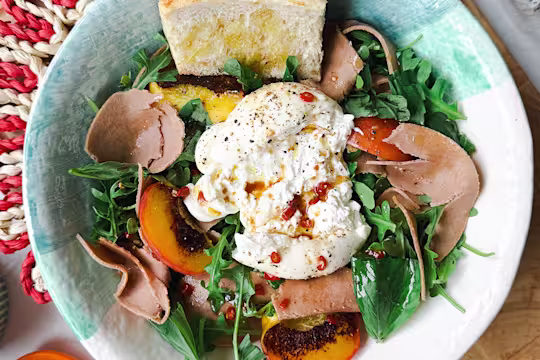 Ceramic bowl on a woven placemat with salad with rocket and basil, burrata cheese, grilled peaches, Quorn Yorkshire Ham Style Slices and toasted bread.