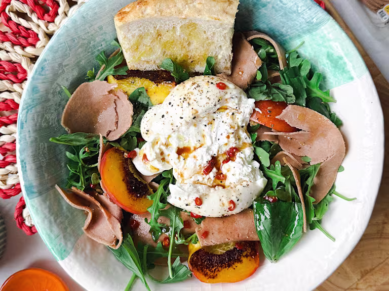 Ceramic bowl on a woven placemat with salad with rocket and basil, burrata cheese, grilled peaches, Quorn Yorkshire Ham Style Slices and toasted bread.