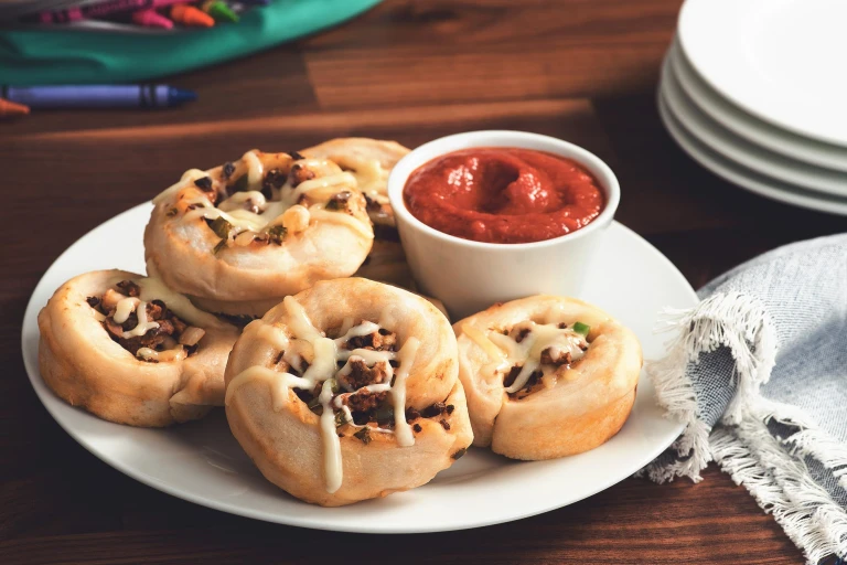 Five pizza roll-ups filled with Quorn Grounds topped with cheese on a white plate with a small ramekin of tomato sauce on the side.