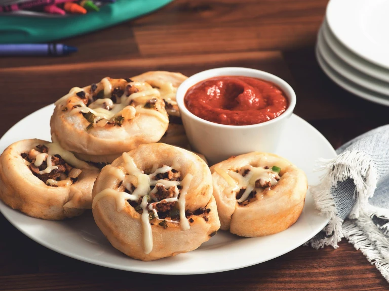 Five pizza roll-ups filled with Quorn Grounds topped with cheese on a white plate with a small ramekin of tomato sauce on the side.