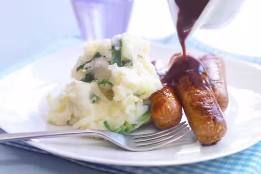Three Quorn Sausages on top of mashed potatoes with gravy poured over them and broccoli on the side.