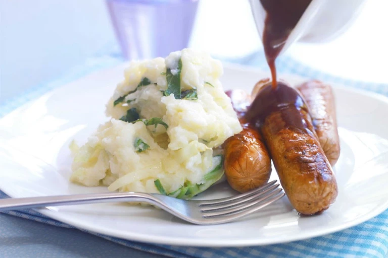 Three Quorn Sausages on top of mashed potatoes with gravy poured over them and broccoli on the side.