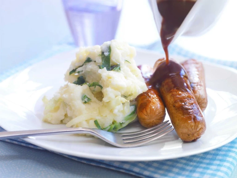 Three Quorn Sausages on top of mashed potatoes with gravy poured over them and broccoli on the side.