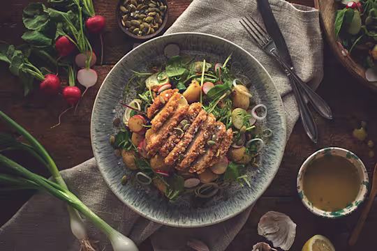 A potato salad with radishes, spring onions, salad mix, pea shoots, and pumpkin seeds topped with a sliced Quorn Southern Fried Burger.