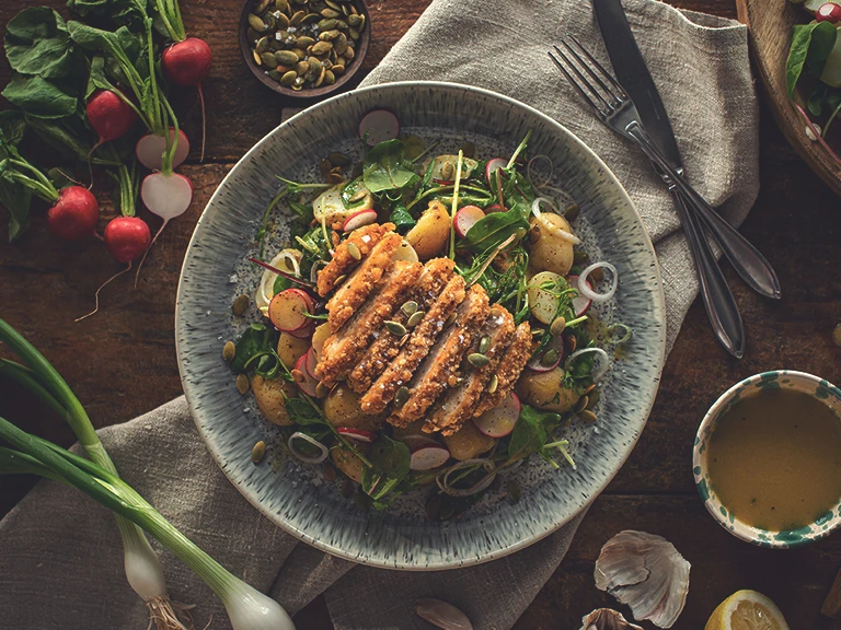 A potato salad with radishes, spring onions, salad mix, pea shoots, and pumpkin seeds topped with a sliced Quorn Southern Fried Burger.