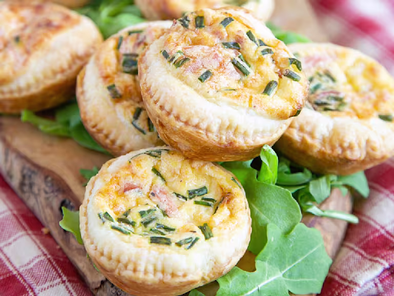 Mini veggie quiches made with Quorn Vegan Ham Slices served on a plate next to tomato slices topped with basil leaves and rocket.