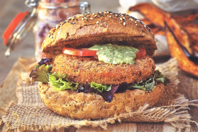 A Quorn Vegan Meatless Spicy Patty on a wholegrain bun with mixed greens, sliced tomatoes, and a coriander and lime dressing with an open jar of slaw in the background.