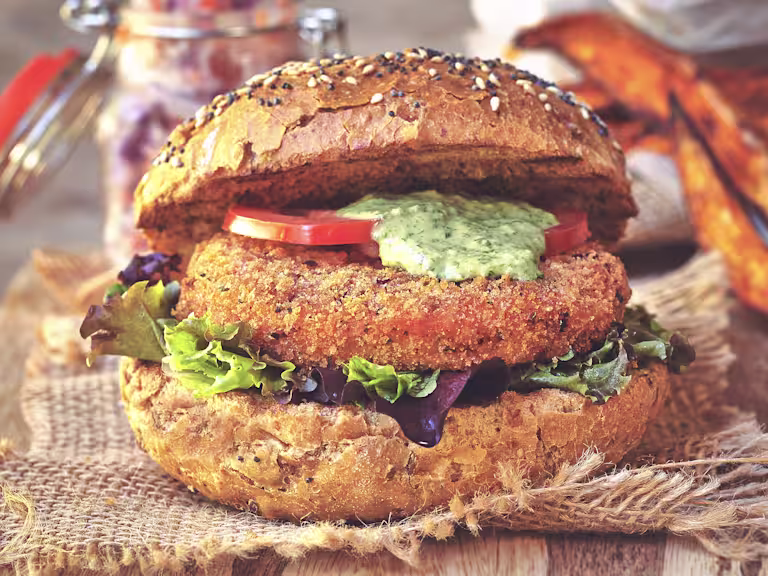 A Quorn Vegan Meatless Spicy Patty on a wholegrain bun with mixed greens, sliced tomatoes, and a coriander and lime dressing with an open jar of slaw in the background.