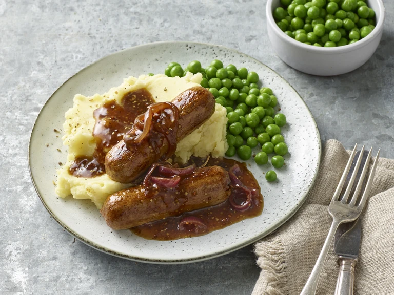 Two Quorn Sausages on top of mashed potatoes with gravy poured over them and peas on the side.