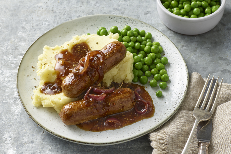 Two Quorn Sausages on top of mashed potatoes with gravy poured over them and peas on the side.