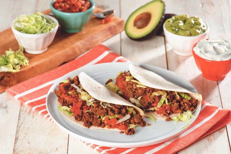 Two flour tortilla tacos filled with Quorn Grounds, shredded lettuce, and diced tomatoes on a white plate surrounded by a variety of toppings.