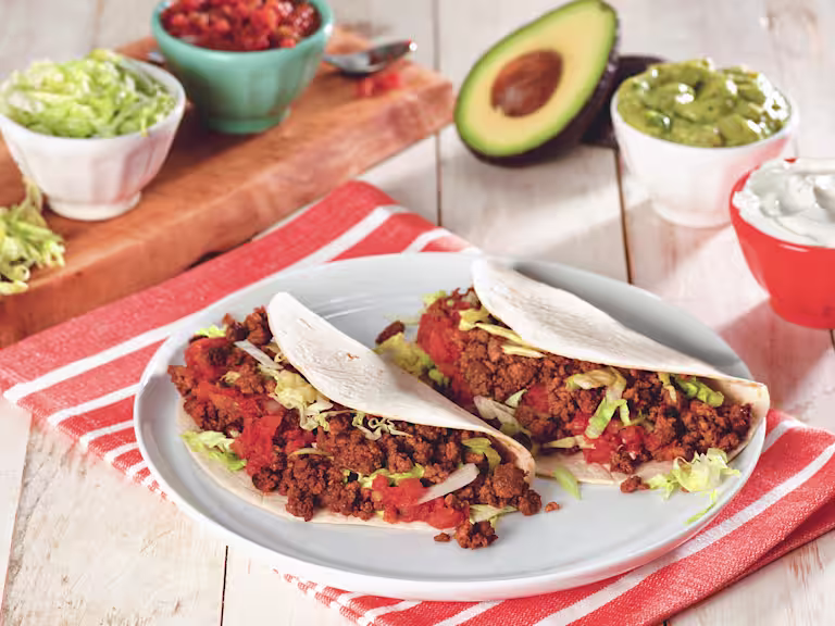Two flour tortilla tacos filled with Quorn Grounds, shredded lettuce, and diced tomatoes on a white plate surrounded by a variety of toppings.