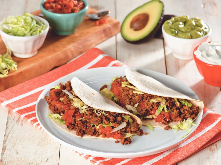 Two flour tortilla tacos filled with Quorn Grounds, shredded lettuce, and diced tomatoes on a white plate surrounded by a variety of toppings.