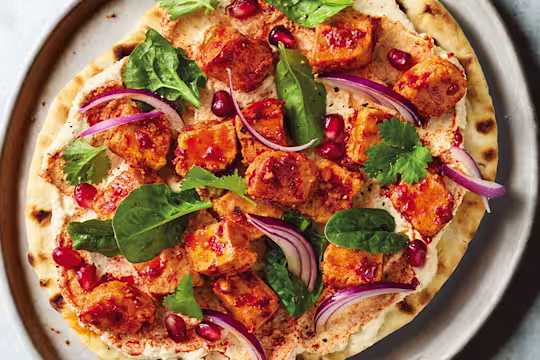Harissa-spiced vegan Quorn pieces on a flatbread with hummus, pomegranate and coriander on a white plate with a blue background.