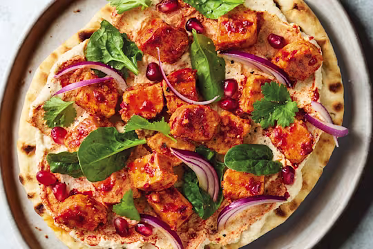 Harissa-spiced vegan Quorn pieces on a flatbread with hummus, pomegranate and coriander on a white plate with a blue background.