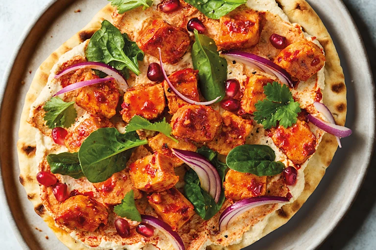 Harissa-spiced vegan Quorn pieces on a flatbread with hummus, pomegranate and coriander on a white plate with a blue background.